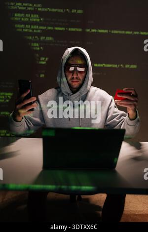Vertical portrait of hooded hacker in glasses sitting at desk, staring at smartphone while holding credit card, laptop open and green code reflecting Stock Photo