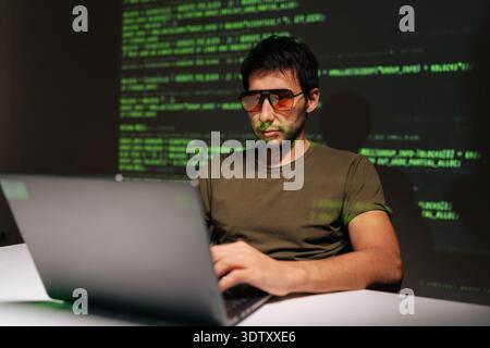 Young man engrossed in coding on laptop, green digital lines of programming code reflecting on face and shirt, indicating intense focus and intricate Stock Photo