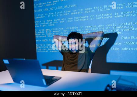 Thoughtful young male developer focused on laptop, debugging code late at night as blue light and projected code lines create cybersecurity and Stock Photo