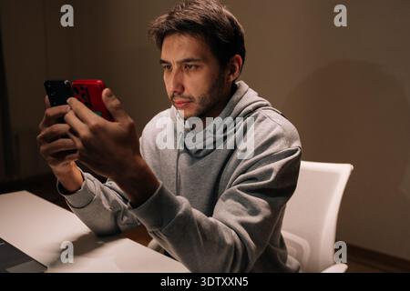 Bearded male in casual hoodie holding two smartphones, focused on comparing apps and messages, multitasking between screens for work or personal Stock Photo