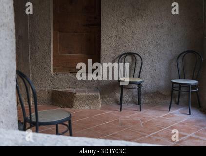 Hree black chairs with woven seats sit on terracotta tile in a rustic room. The textured wall and wooden shuttered window give a cozy, unfinished appe Stock Photo