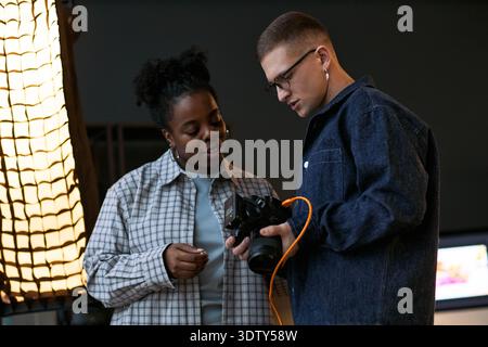 Young Black woman and young Caucasian man examining digital camera together in studio setting, both standing close and focusing on camera screen, professional lighting equipment visible Stock Photo