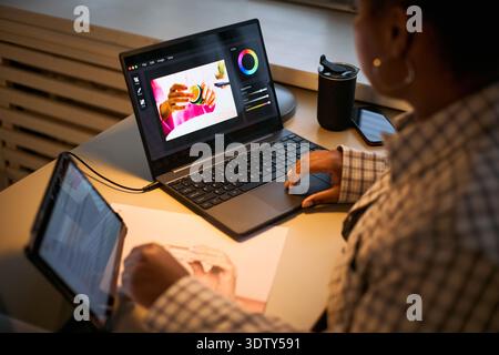Black woman using laptop and tablet while editing photo, sitting at desk with stylus in hand, focusing on creative project, technology and creativity concept Stock Photo