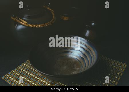 A Japanese ceramic bowl and teapot set displayed on a bamboo mat under moody lighting. The setup captures traditional Japanese tableware elegance. Stock Photo