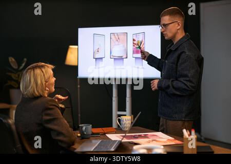 Young Caucasian man presenting digital marketing strategy to middle aged Caucasian woman, using large screen with product images in modern office setting during business meeting Stock Photo