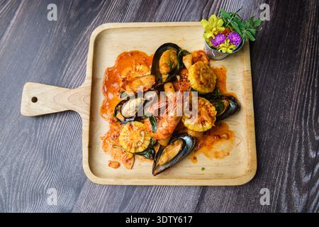 Top-down view of spicy stir-fried seafood with shrimp, mussels, and baby corn, elegantly served on a wooden plate with delicate flower garnish. Stock Photo