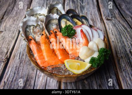 A rustic fresh seafood platter of oysters, shrimp, mussels, scallops, and squid served on ice with lemon and parsley, placed on a wooden surface. Stock Photo