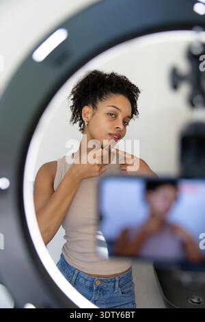 Woman in beige top pointing chin, holding green tool, recording on phone tripod with ring light Stock Photo