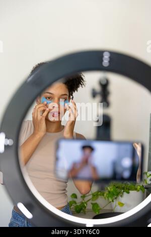 Woman applying blue undereye hydrogel patches at vanity mirror, filming on phone with ring light Stock Photo