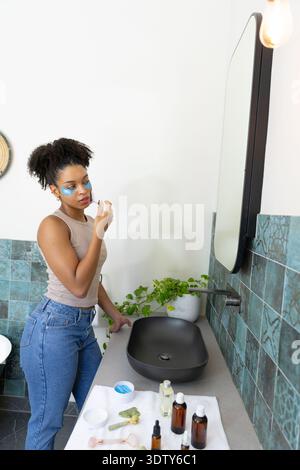 African American woman using skincare, wearing under-eye patches at bathroom sink, copy space Stock Photo