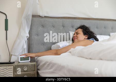 Woman reclining on bed reaching left arm toward white digital alarm clock on nightstand Stock Photo