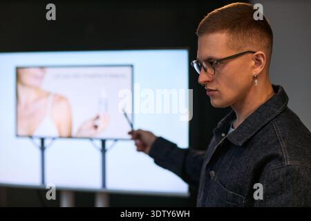 Young adult Caucasian man presenting information on digital screen, holding pointer and gesturing toward image of woman with cosmetic product during business presentation Stock Photo