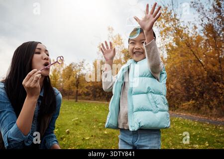 Asian, daughter or mom blowing with bubbles in park for playful childhood or outdoor bonding. Mother, child or kid with wand for parenting Stock Photo