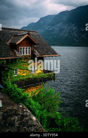 Charming house covered in ivy perched on a cliff overlooking a dark, moody lake and mountains Stock Photo