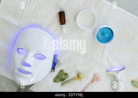 White LED mask is glowing on terry towel, amber dropper and blue eye patches are resting Stock Photo