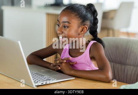 Silver laptop is sitting open on wooden dining table near kitchen island with soft natural light Stock Photo