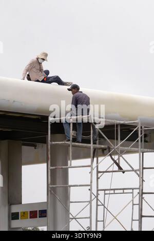 Two construction workers sitting on scaffolding and pipeline structure during industrial maintenance work at an outdoor job site in Cambodia. Stock Photo