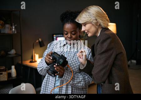 Young adult Black woman showing digital camera screen to mature Caucasian woman, both standing close together and examining captured images in modern workspace setting Stock Photo