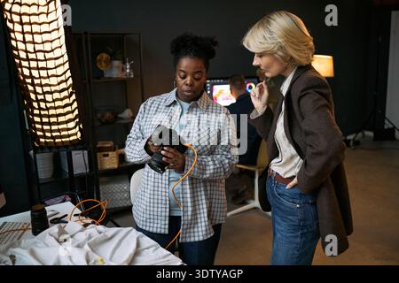 Black woman and Caucasian woman reviewing images on digital camera during professional photo shoot, standing near table with fabric and photography equipment in studio setting Stock Photo