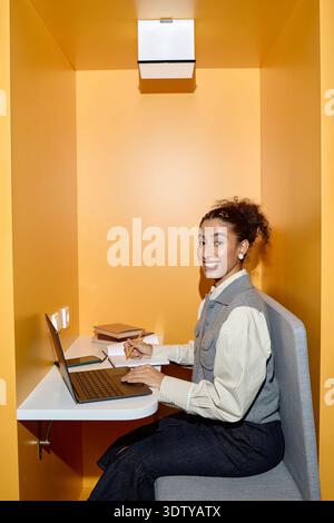 Young Adult Multiethnic Woman Using Laptop and Writing Notes in Library Study Booth Stock Photo
