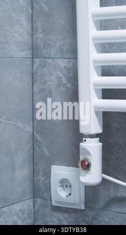 White towel radiator installed on gray tiles with electrical outlet visible nearby Stock Photo