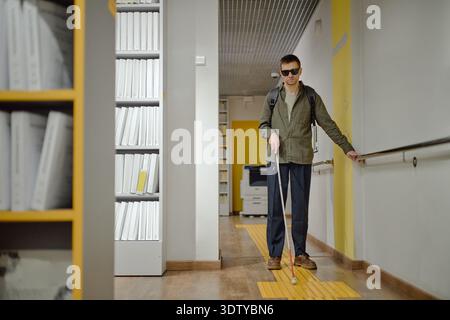 Young Adult Man Navigating Library Hallway Using Cane and Rail Stock Photo