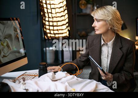 Caucasian middle aged woman working at desk using desktop computer and holding digital tablet, focusing on screen with professional lighting equipment in background, engaging in creative project Stock Photo