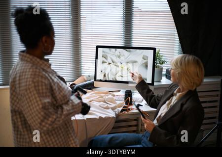 Caucasian mature woman sitting and pointing at computer monitor while Black woman standing nearby holding camera, both collaborating on creative project in modern workspace Stock Photo