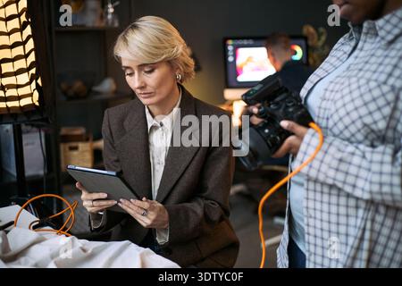 Caucasian middle aged woman using digital tablet while Black woman holding professional camera, both working together in creative studio with man visible in background operating computer Stock Photo