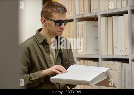 Young Man Reading Braille Book in Library While Using Cane Stock Photo