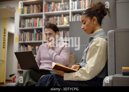 Young Man Using Laptop While Young Black Woman Reading Book in Library Stock Photo