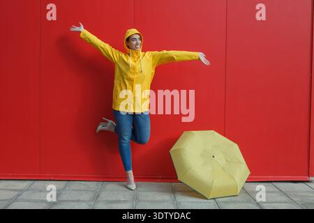 A woman wearing a yellow rain jacket and high heels stands on one leg, smiling and spreading her arms in front of a bright red wall. Stock Photo