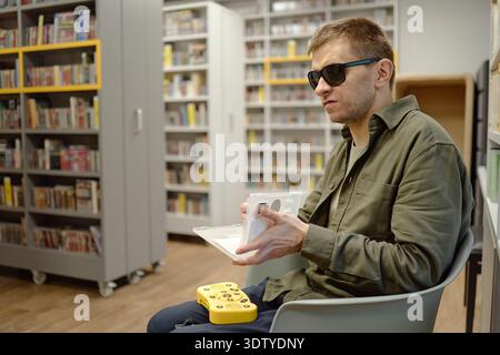 Young Man Reading Braille Book with Assistive Device in Library Stock Photo