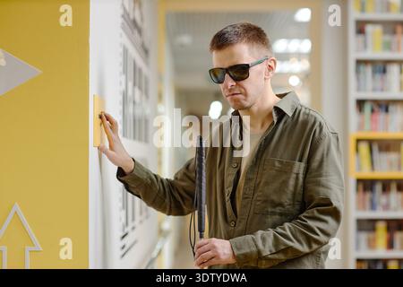 Young Man Navigating Library with Cane and Touching Tactile Sign Stock Photo
