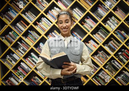Young Adult Multiethnic Woman Smiling While Holding Book in Library Setting Stock Photo