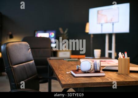 Modern office workspace featuring empty chair and wooden desk with closed laptop, wireless headphones, notebook, and stationery, large monitor screen visible in background Stock Photo
