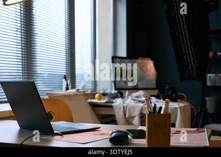 Modern workspace featuring open laptop on desk with wireless mouse and pencil holder, creative studio environment visible in background with blurred art supplies and large window blinds Stock Photo