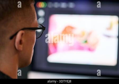 Young adult Caucasian man wearing glasses sitting in front of computer monitor working on digital project, viewing blurred image of hands holding object on screen Stock Photo
