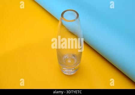 A single empty glass vase is centrally placed on a split background of bright yellow and light blue paper Stock Photo