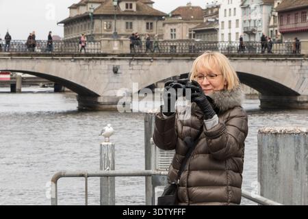 Blonde attractive woman in modern glasses making photo on camera against city bridge in cloudy winter day. Europe cityscape. Stock Photo