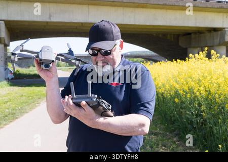 Man in blue navy shirt holding drone and controller in his hands before the start of the flight against the background of field with rapeseed Stock Photo