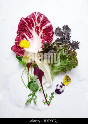 A collection of different lettuce leaves and edible flowers is arranged on a white marble countertop. These ingredients are ideal for making a fresh v Stock Photo