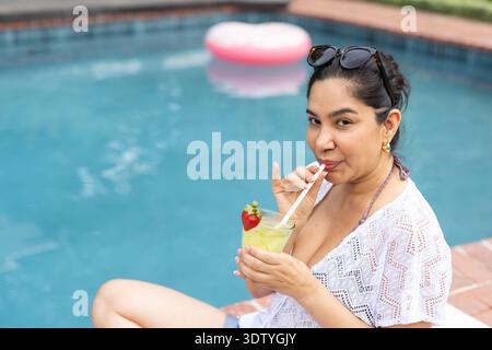 Woman sitting on right pool edge wearing cover-up, sipping cocktail, sunglasses on head, copy space Stock Photo