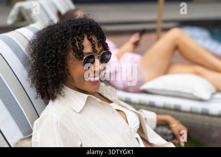 Diverse female friends reclining on striped chaise at poolside, wearing sunglasses, holding phone Stock Photo
