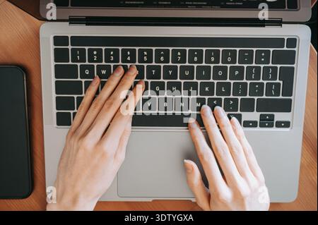 Overhead view of hands typing on a laptop keyboard placed on a wooden desk with a smartphone nearby. Modern workspace scene illustrating remote work, Stock Photo