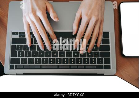 Overhead close-up of female hands typing on a laptop keyboard at a wooden desk with smartphone beside. Modern workspace concept ideal for business, re Stock Photo
