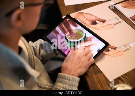 Young adult man using tablet, viewing close up image of hands applying skincare cream, sitting at table with printed skincare product photos, engaging in product research Stock Photo