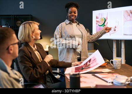 Black young adult woman presenting skincare product images on digital screen to Caucasian man and woman during creative team meeting, colleagues discussing project Stock Photo