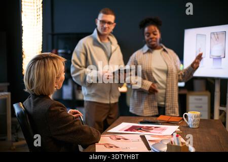 Caucasian middle aged woman sitting at desk, listening to young adult Caucasian man and young adult Black woman standing and discussing design displayed on monitor Stock Photo