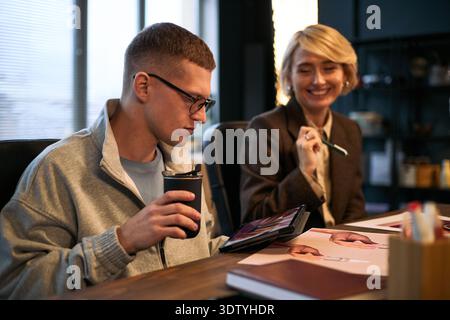 Caucasian young adult man holding reusable coffee cup and using digital tablet while sitting at desk next to smiling Caucasian middle aged woman, discussing creative project Stock Photo